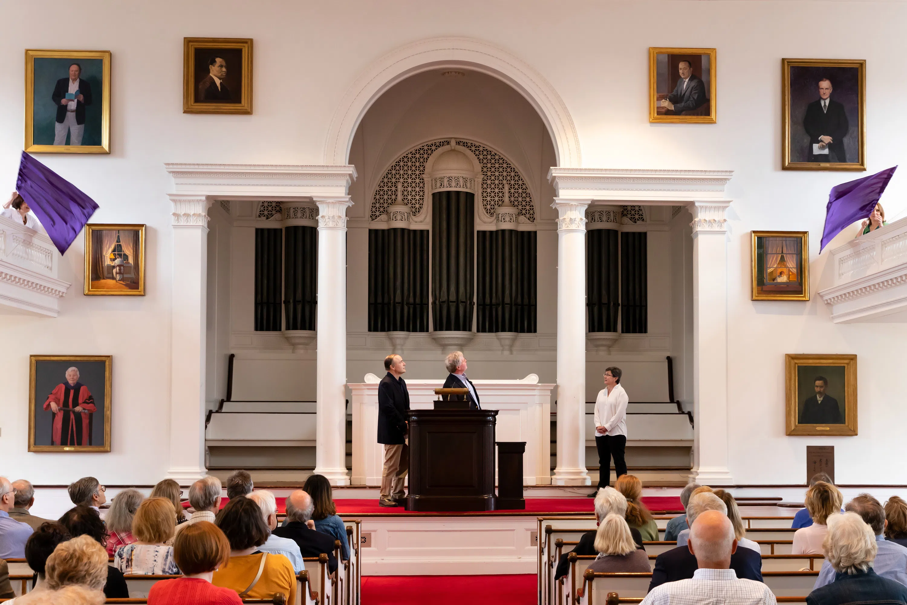 Interior shot of Johnson Chapel with two men on stage and crowd looking on as paintings on wall are unveiled.
