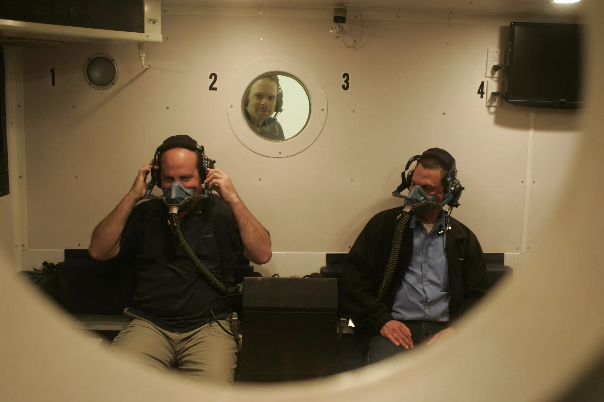 Two men inside altitude chamber with oxygen masks on.
