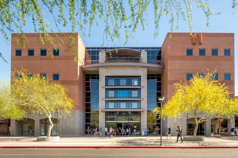 Four-story brick and glass building with students walking out of the entrance and two palo verde trees in yellow bloom in front of building.