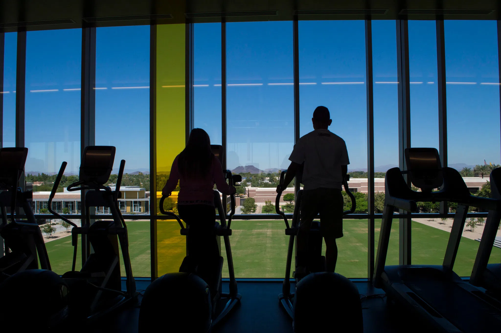 Two students work out on eliptical machines while looking out the window of a view of West campus.