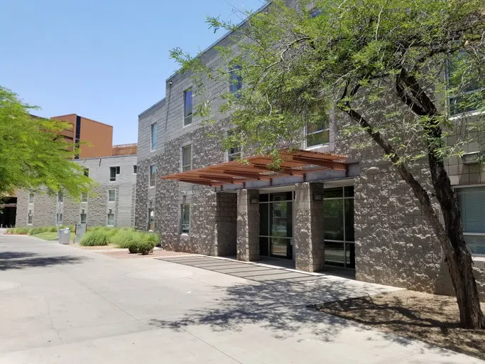 A palo verde tree provides shade near a covered entrance to a three-story cinderblock building. 