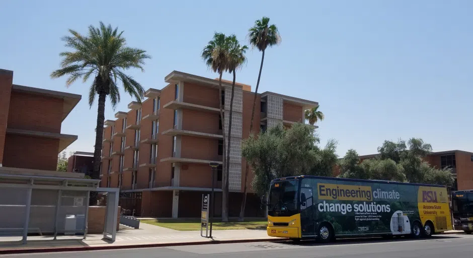 A bus with an ASU ad on the side saying "Engineering climate change solutions" sits in front of a five-story brick building with concrete awnings over every window. 