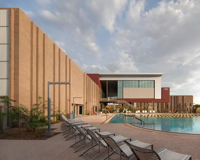 Lounge chairs lined up around pool with fitness complex to the left. 