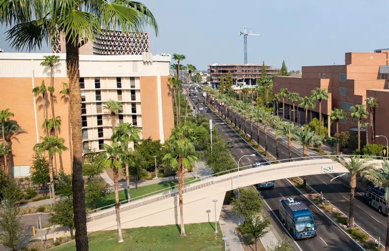 A pedestrian bridge spans a four-lane road in front of seven-story brick building.