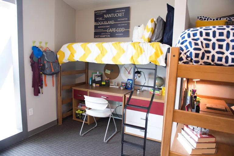 A room with a desk and chair underneath a lofted bed with yellow and white zigzag striped bedspread. To the right of the bed is another lofted bed with desk underneath. 