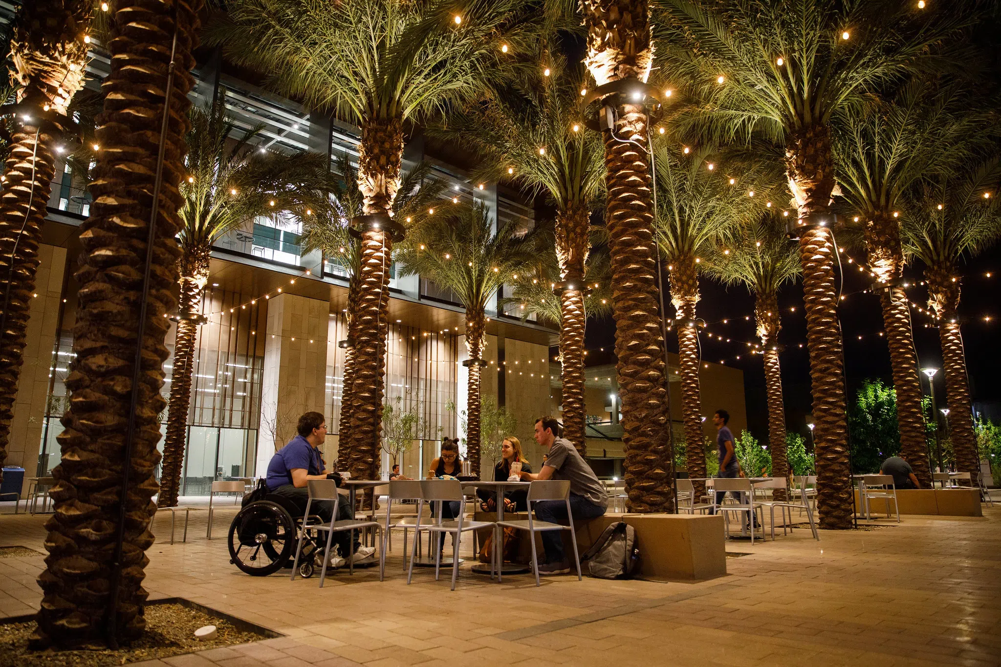 Student a wheelchair sits at a table with three other students outside the Student Pavilion at night. They are surrounded by palm trees with strings of lights hanging from the trees. 