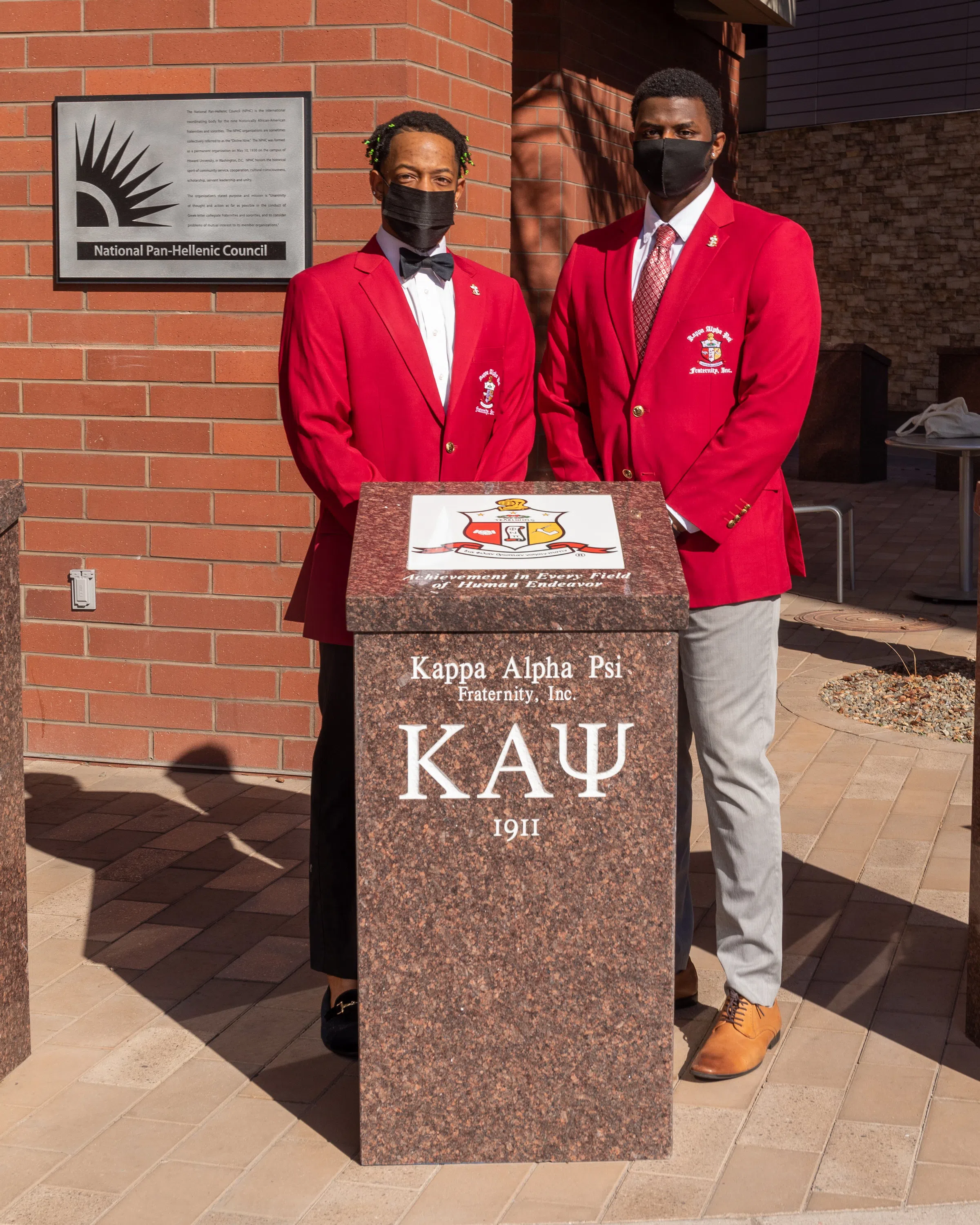 Two representatives from the Kappa Alpha Psi fraternity stand next to their plaque wearing their fraternity's red blazers. 