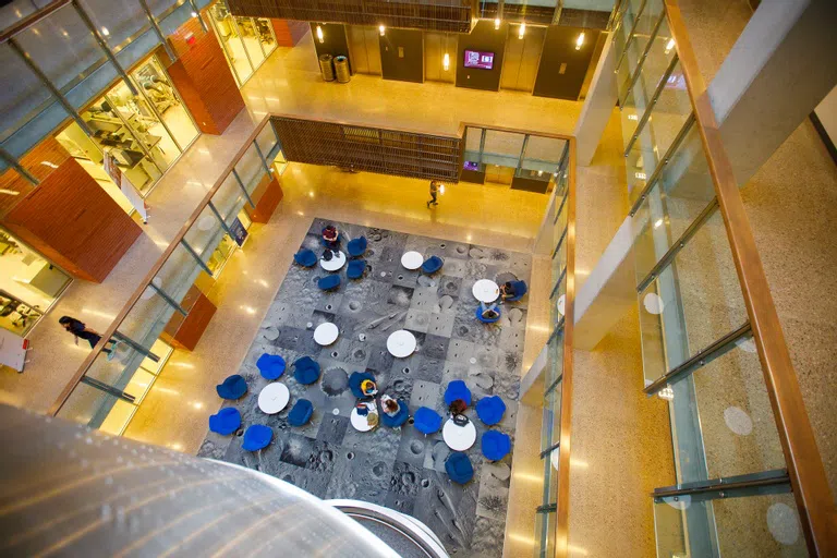  View from top of a four-story atrium looking down to people working at white tables and blue chairs on top of carpet with images of moon craters on it. 