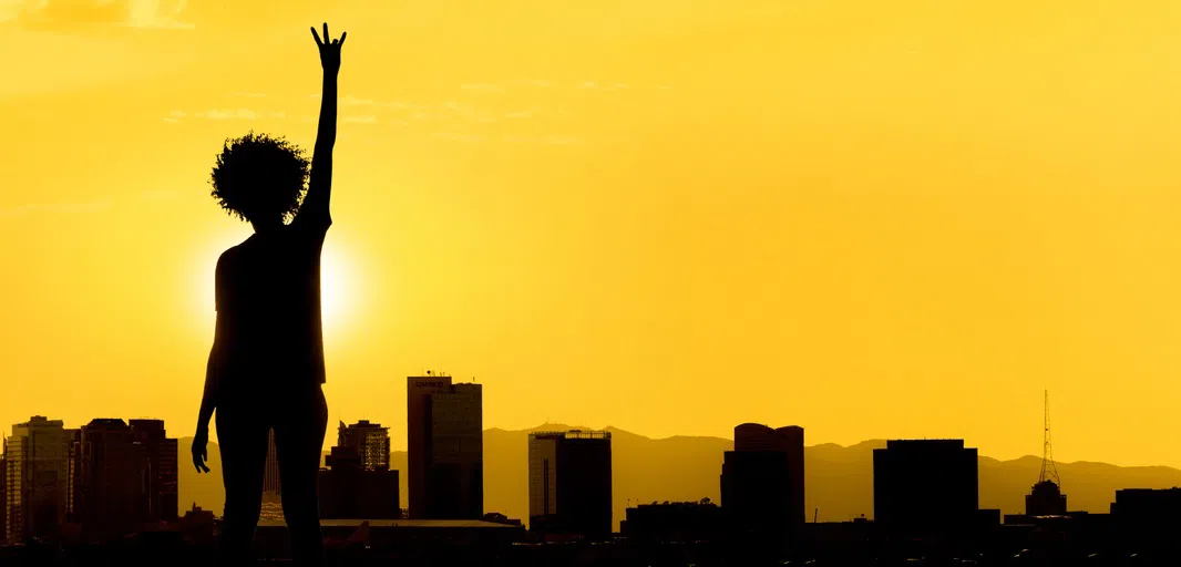 woman in silhouette holding up her right hand in ASU pitchfork hand sign, with the Phoenix skyline in silhouette in front of her and a golden sunset sky. 