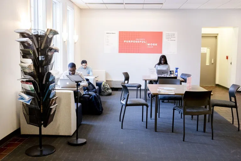 Three students sit behind laptops at separate tables in front of a banner that reads 'Purposeful Work'