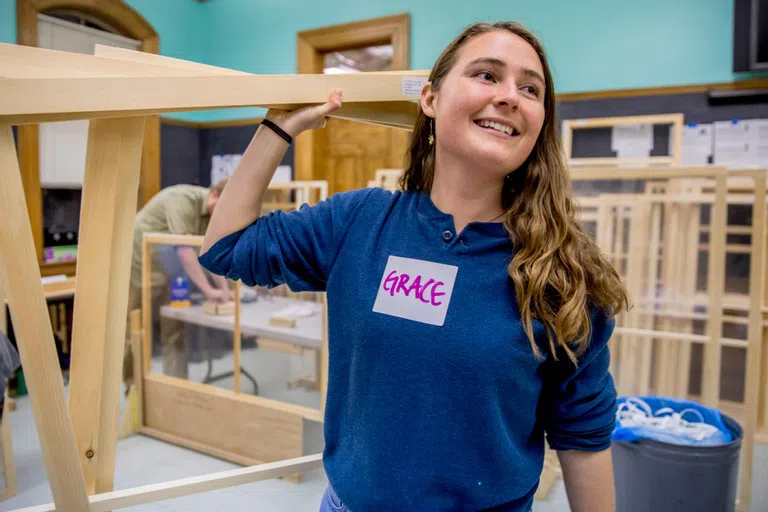 A student with a nametag that reads 'Grace' carries a beam of wood