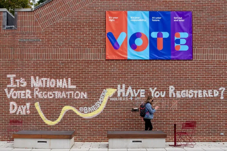 A student walks past a large chalk mural on a brick building that asks 'Have you registered [to vote]?'