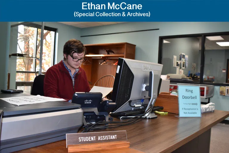 Male student sits at desk with computer in library. A sign reads "Student Assistant"