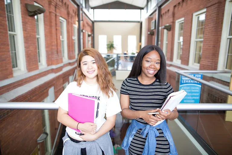 Two smiling students holding folders in a hallway