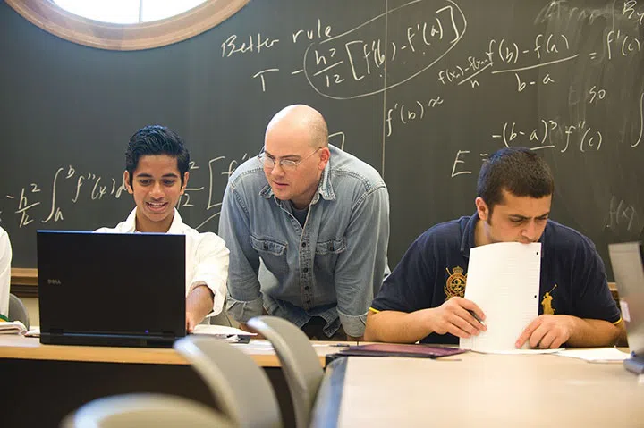 Students and a professor look at a laptop. Math formulas are on the board behind them.