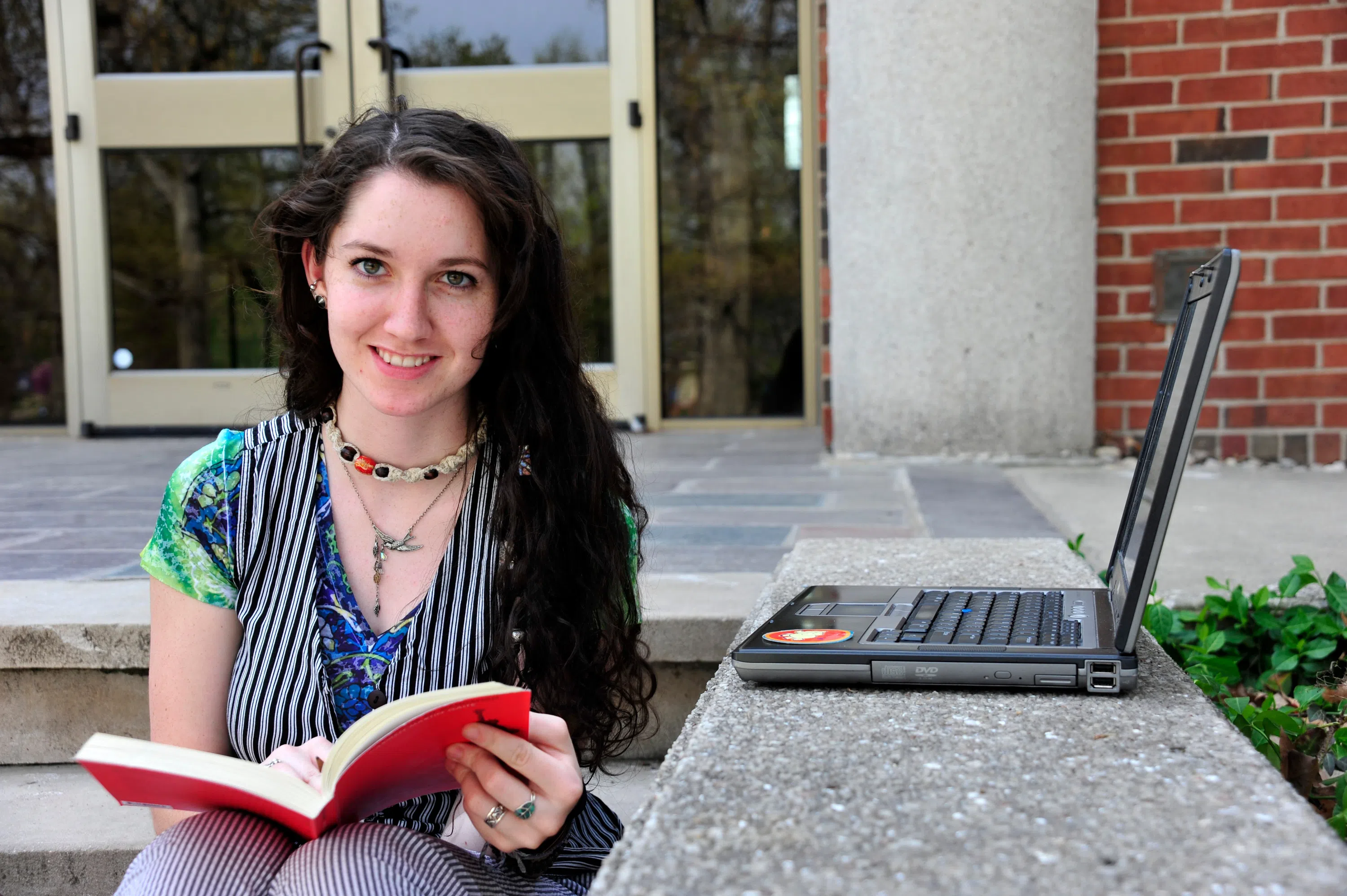 Students sits on steps of library holding a red notebook. Beside her is an open laptop 