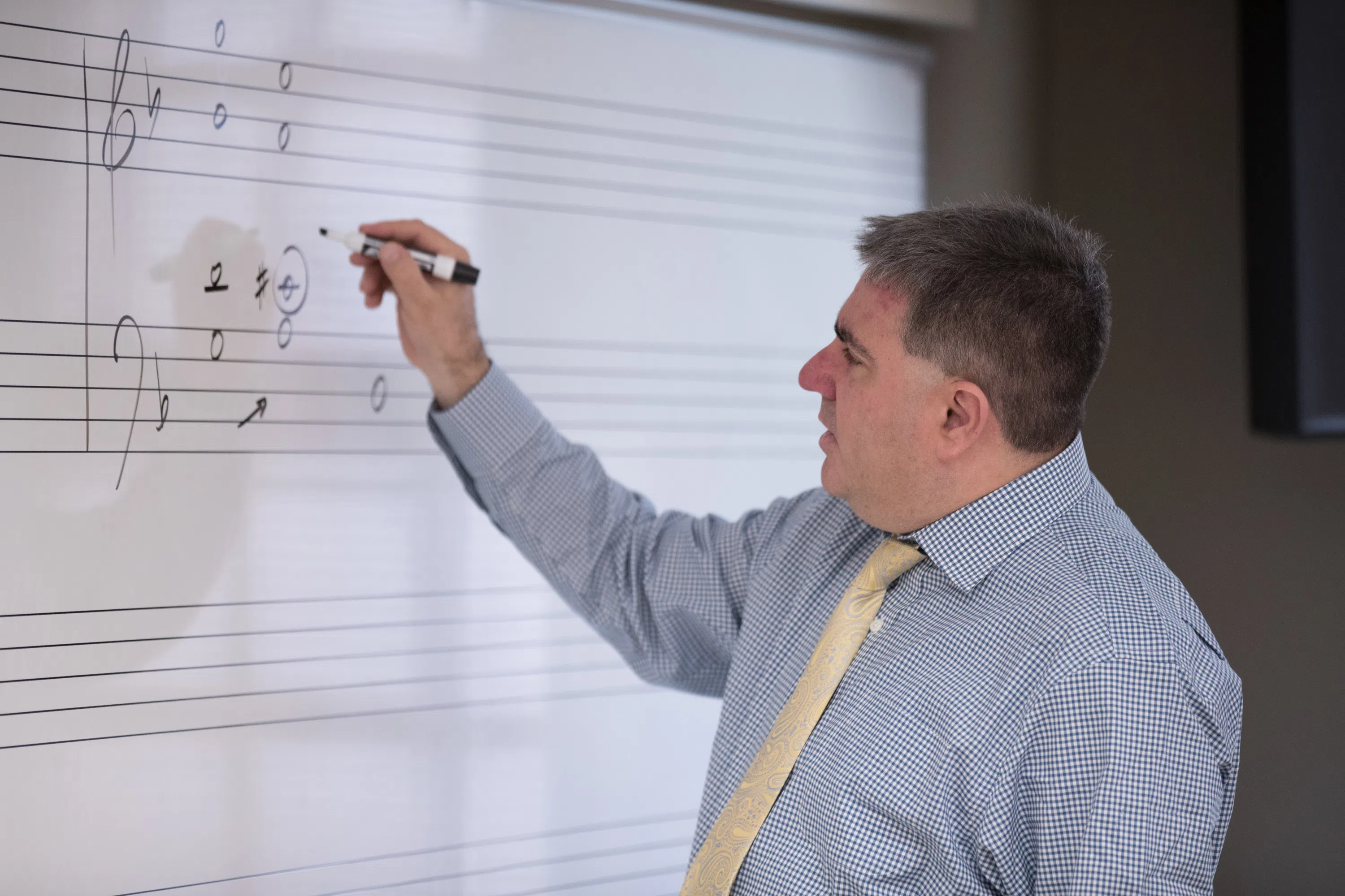 Professor stands at white board with music bars writing music notes with a black marker.