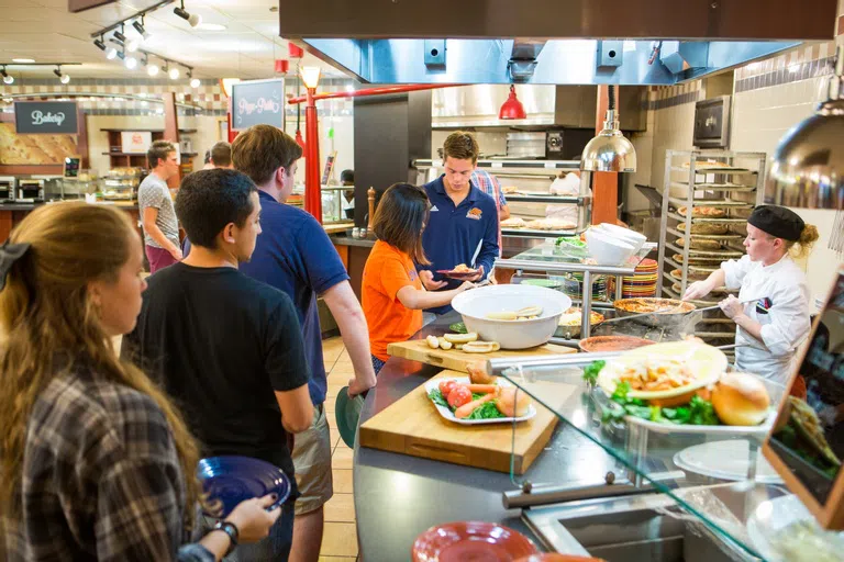 5 students line up to get food from the pasta station, with a female food service worker working on the other side of the counter