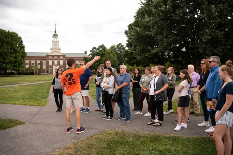 Male tour guide points off screen for tour group, with bertrand library behind them