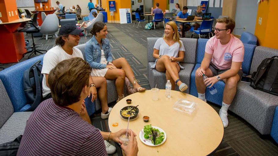 a group of students in comfortable chairs circling around a round table, with people studying in the large space behind them