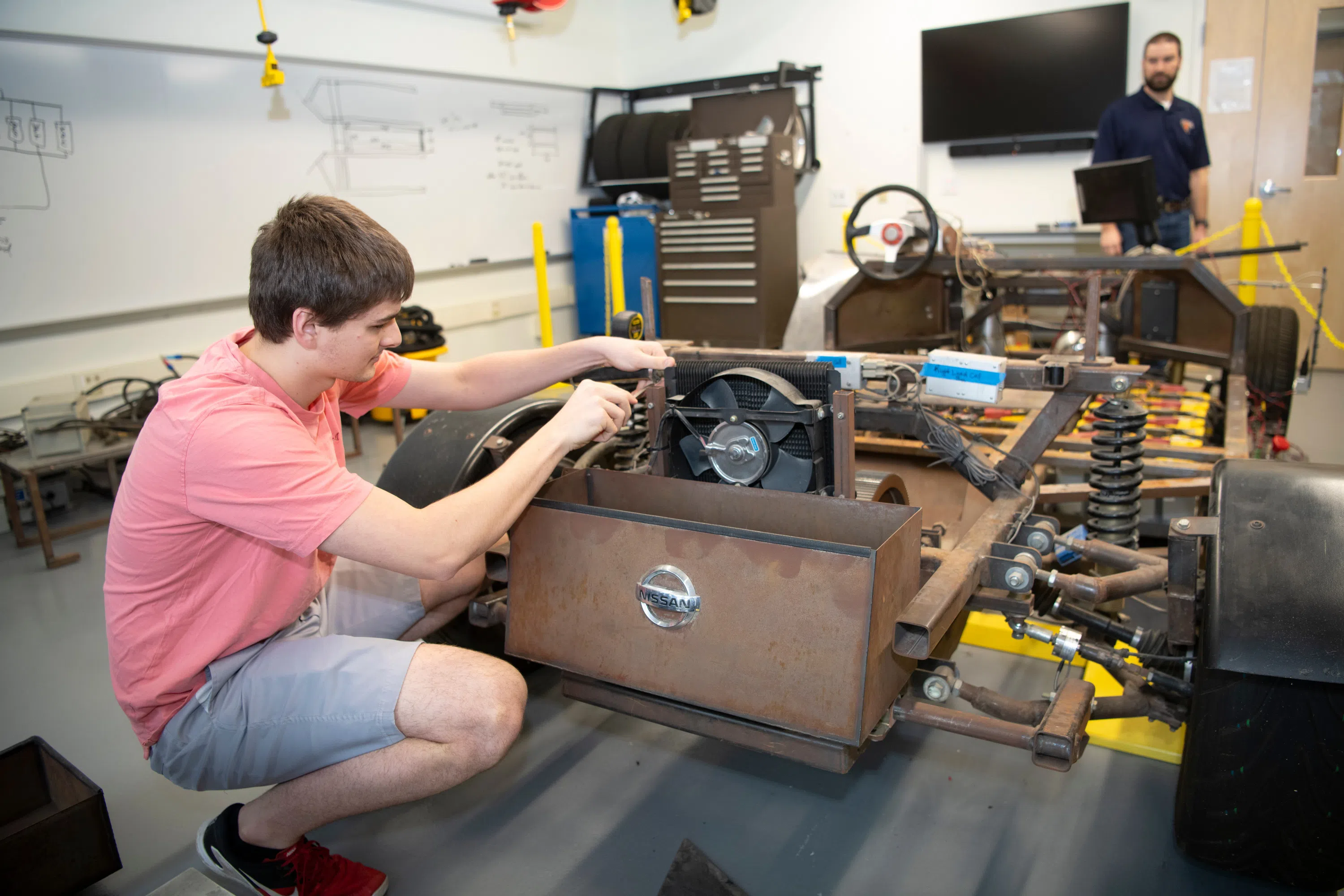 a student works on a home-built car, a member of faculty in the background