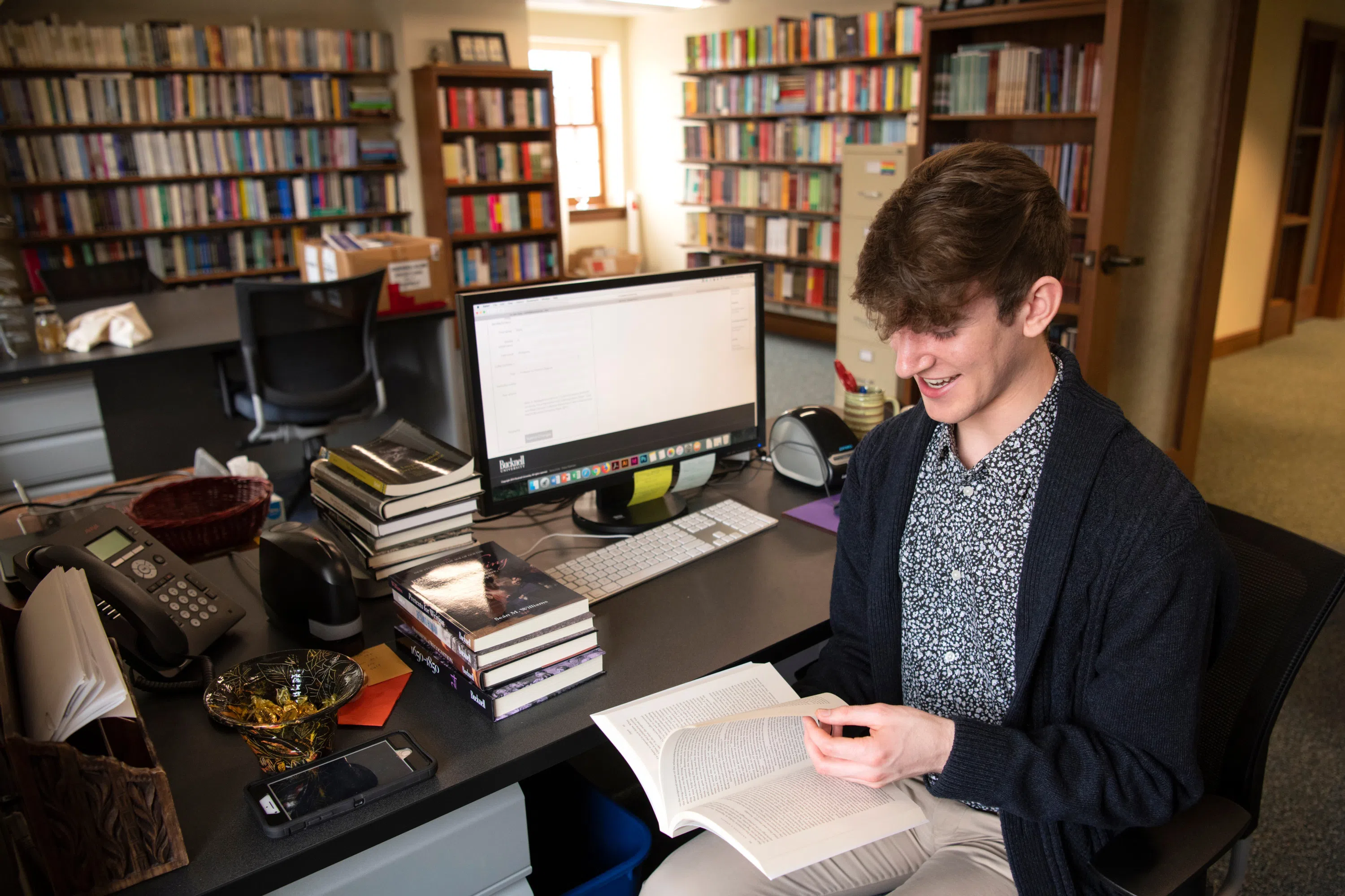 a male student looks through a book with a computer next to him, the room filled with books on bookshelves in the background