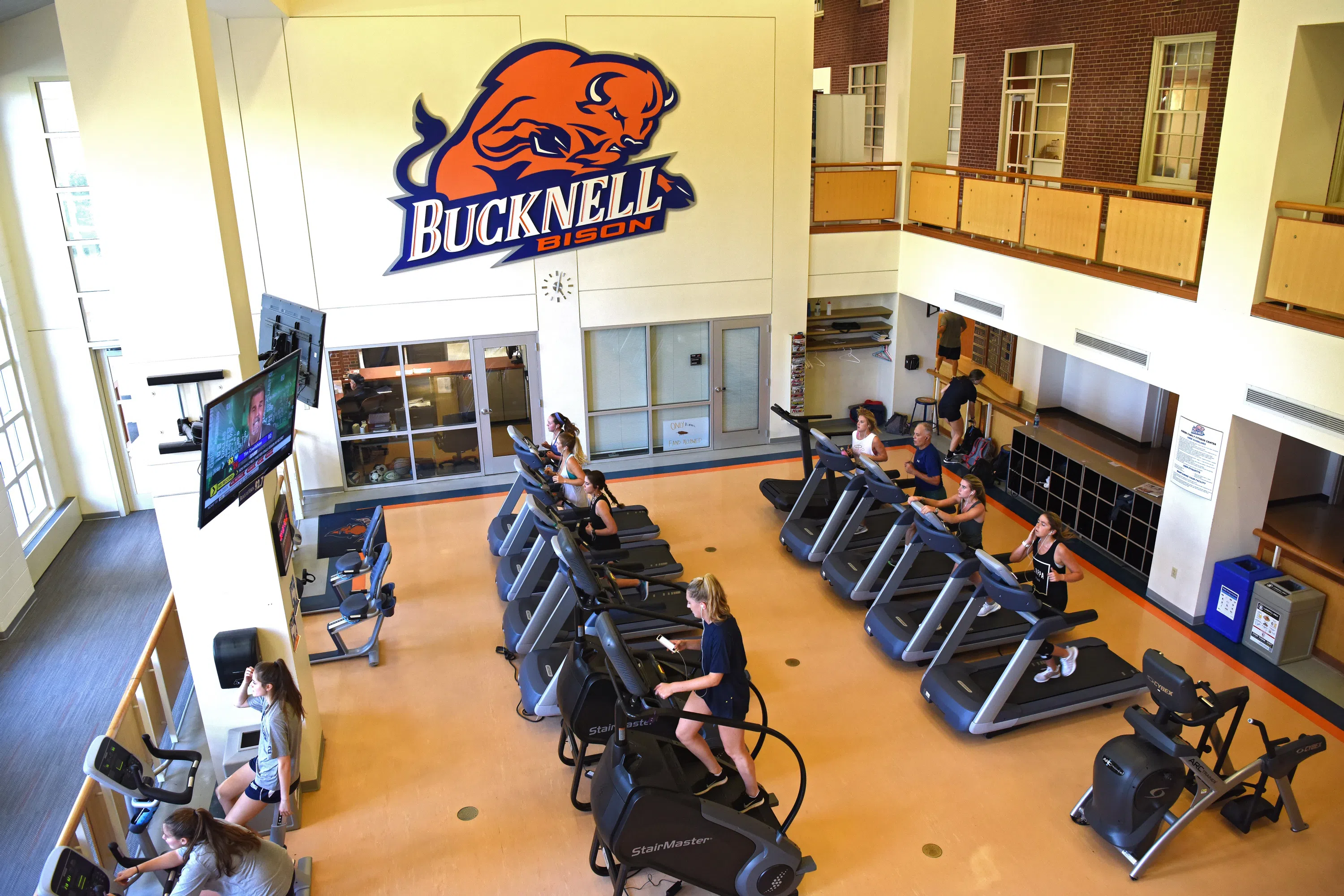 people use exercise equipment in the gym, with a large Bucknell Bison logo on the wall in the background