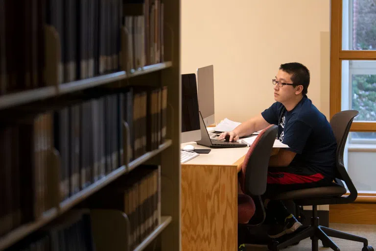 A student works on a laptop at a desk in the background, books out of focus in the foreground