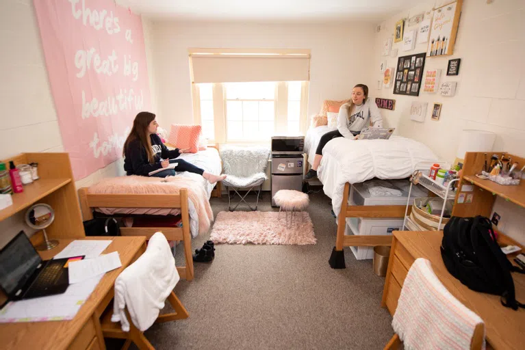 two female students sit on beds on opposite ends of a beautifully decorated dorm room