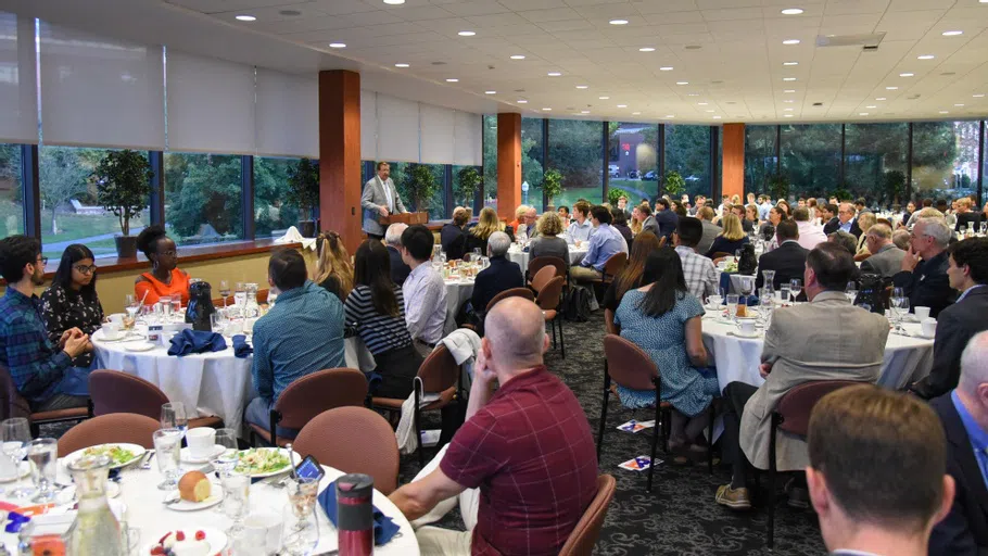 a large group of people fill a banquet hall listening to President Bravman speaking at a podium in the background