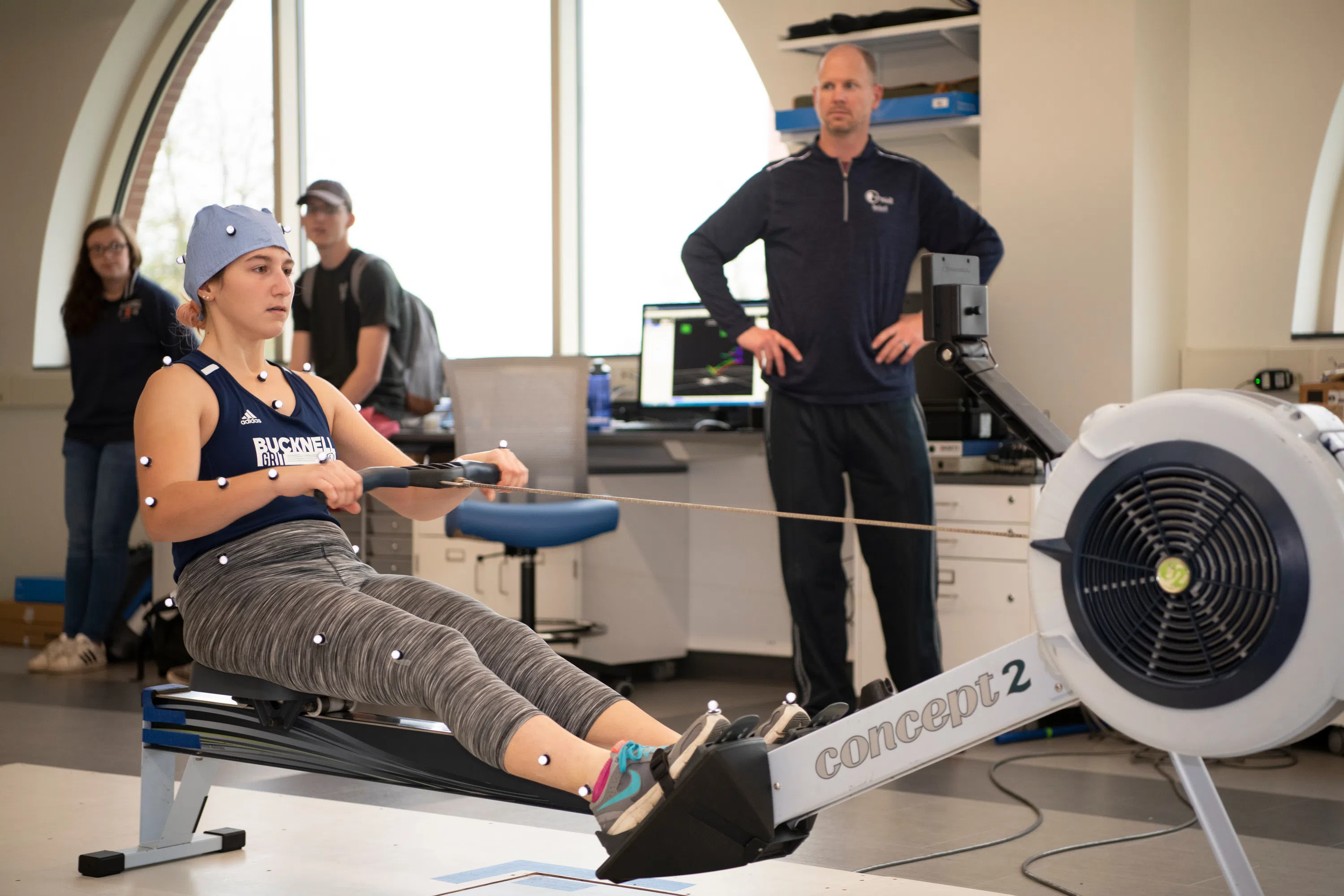 a female student covered in motion capture markers uses a rowing machine in the motion capture lab, with faculty and students looking on