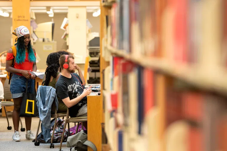 Books out of focus in the foreground, a student listening to headphones sitting at a table in the background, two students talking behind him