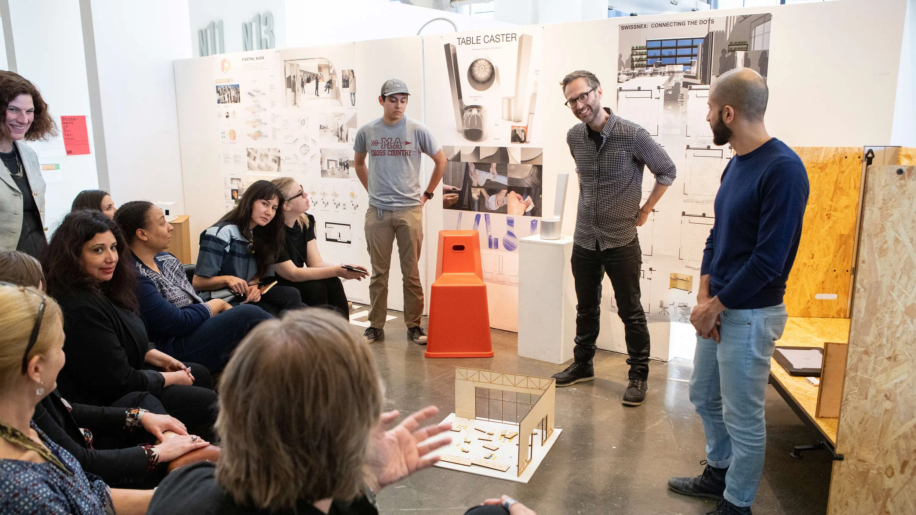 Three Interior Design students cluster around a small plywood model on the floor; classmates and panelists form a half circle around them and critique their work.