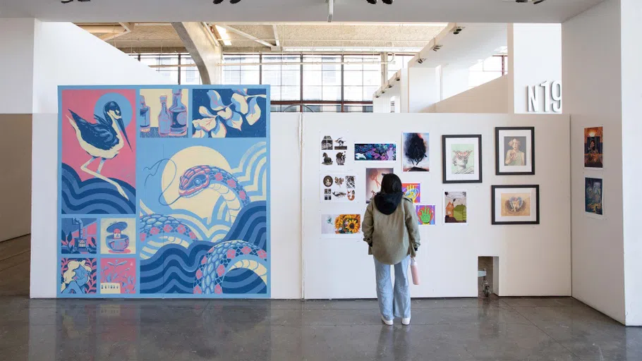 A student stands in front of a while wall displaying 14 works of art and a large mural.