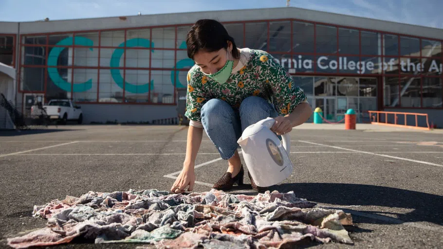 A student crouches down in the Backlot, pouring hot water onto a painting on the ground.