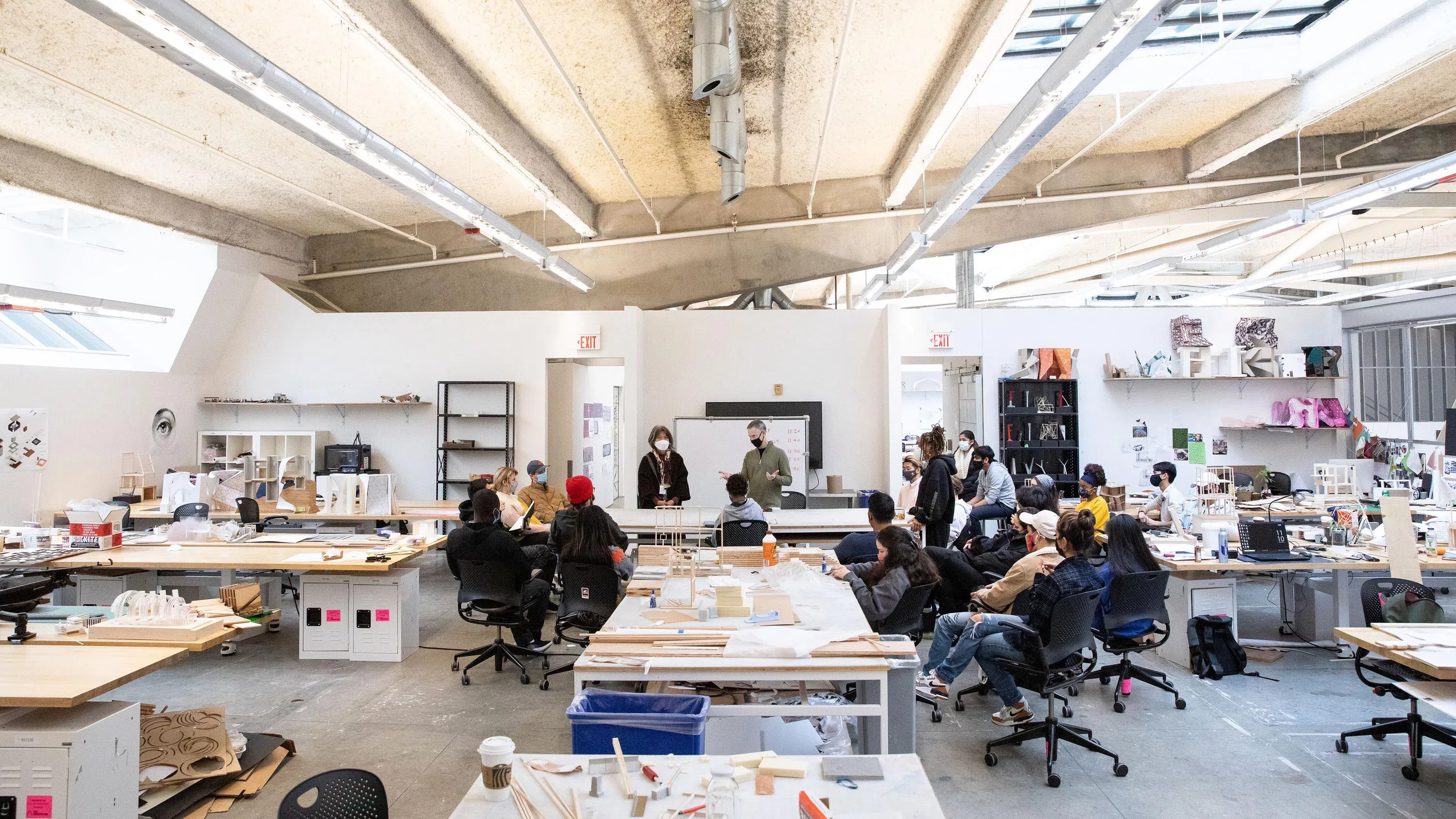 A wide shot of students seated in the Architecture studio, listening to two professors lecture.