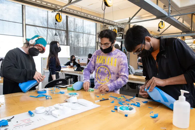 Three students work at a wooden table, adding blue tape to plastic shoe molds.