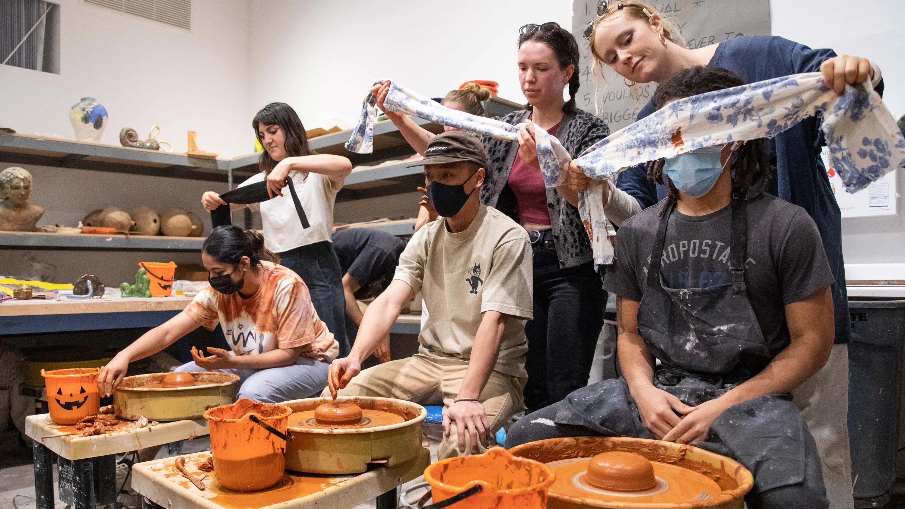 Three students standing up put blindfolds on three students sitting at a potter’s wheel