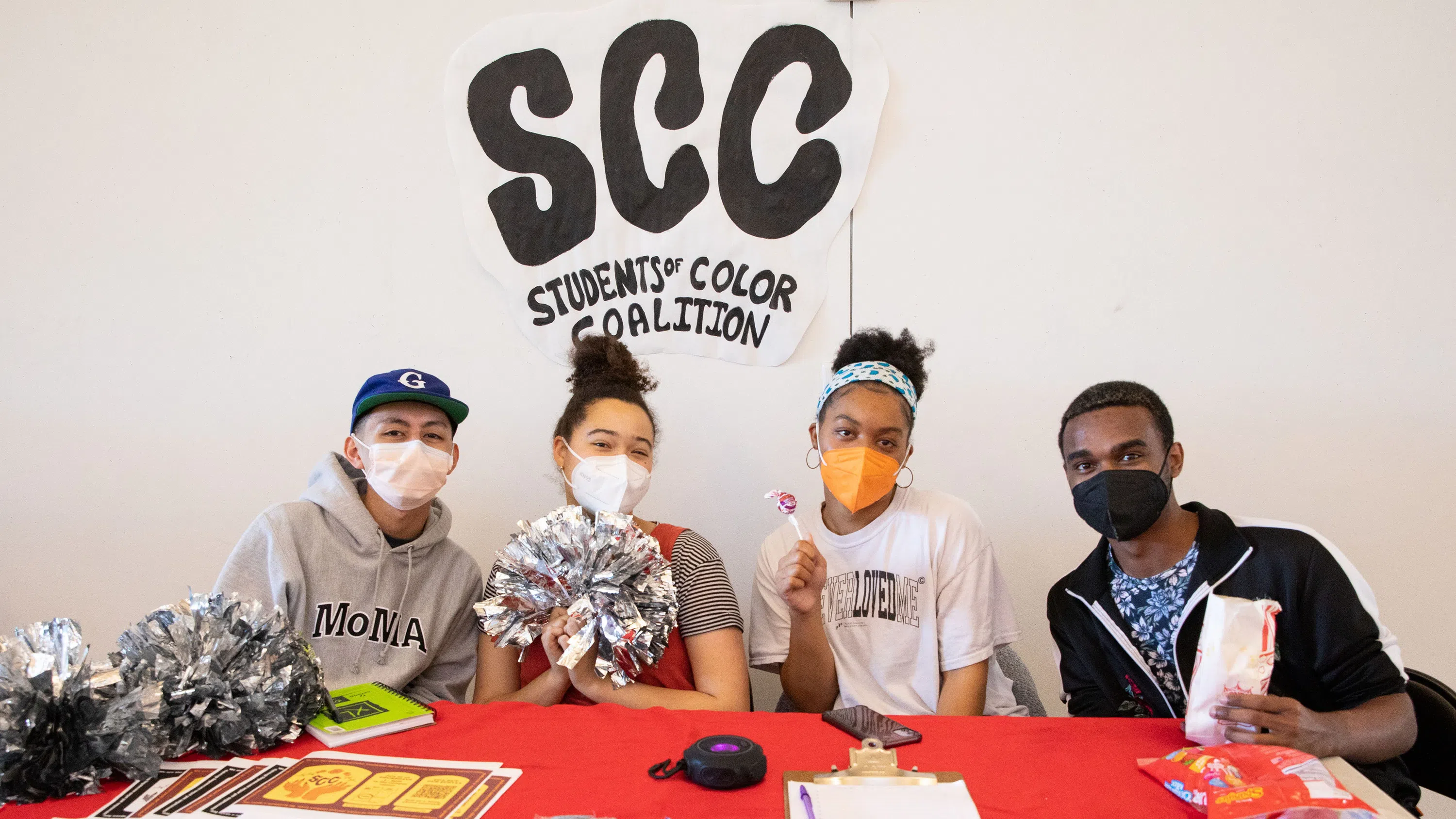 Four masked students sit at a table with a red tablecloth, candy, and collateral. Large letters on the wall behind them read SCC: Students of Color Coalition.