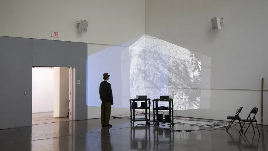 A person views a layered projection on a white wall in the Nave Alcove. Large-scale paintings are on the floor and chairs surround the installation.