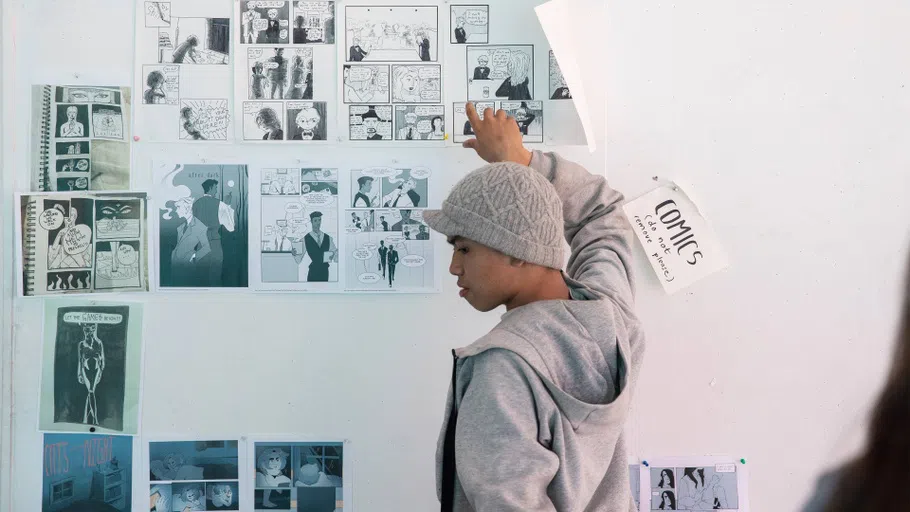 A student pins up black-and-white comic strips on a white wall.