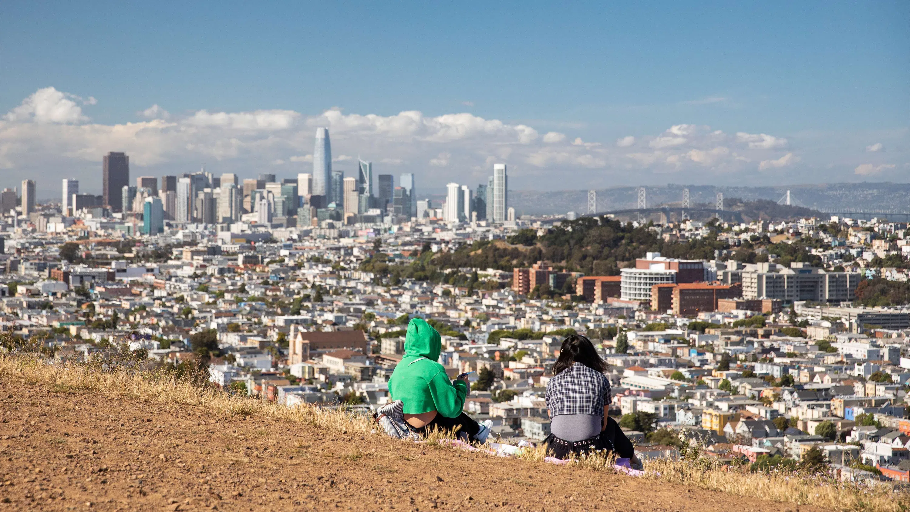 Two students sit on a slope looking at the SF skyline and Bay Bridge.