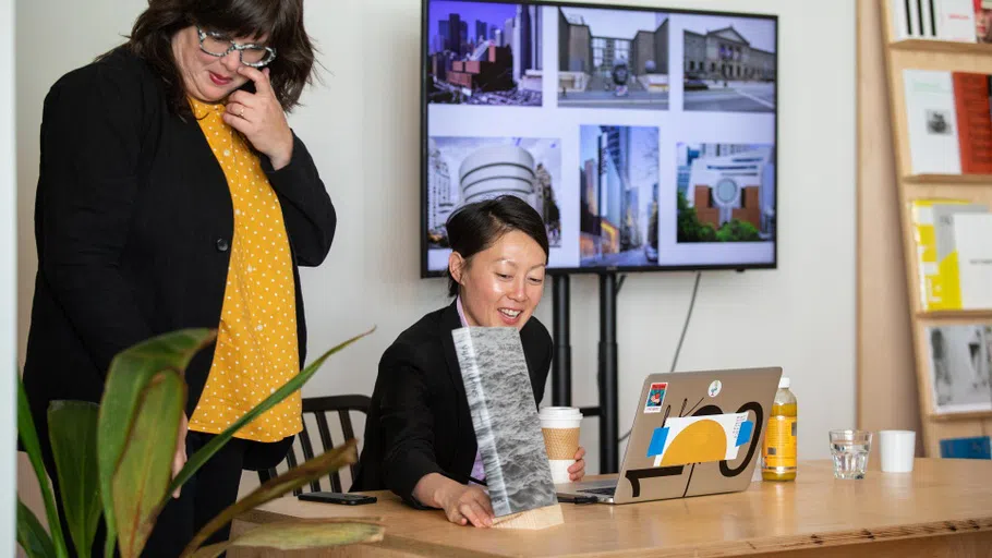 Curator Nancy Lim sits at a desk and points to a recent catalog about Vija Celmins; former faculty Christina Linden stands to her left.