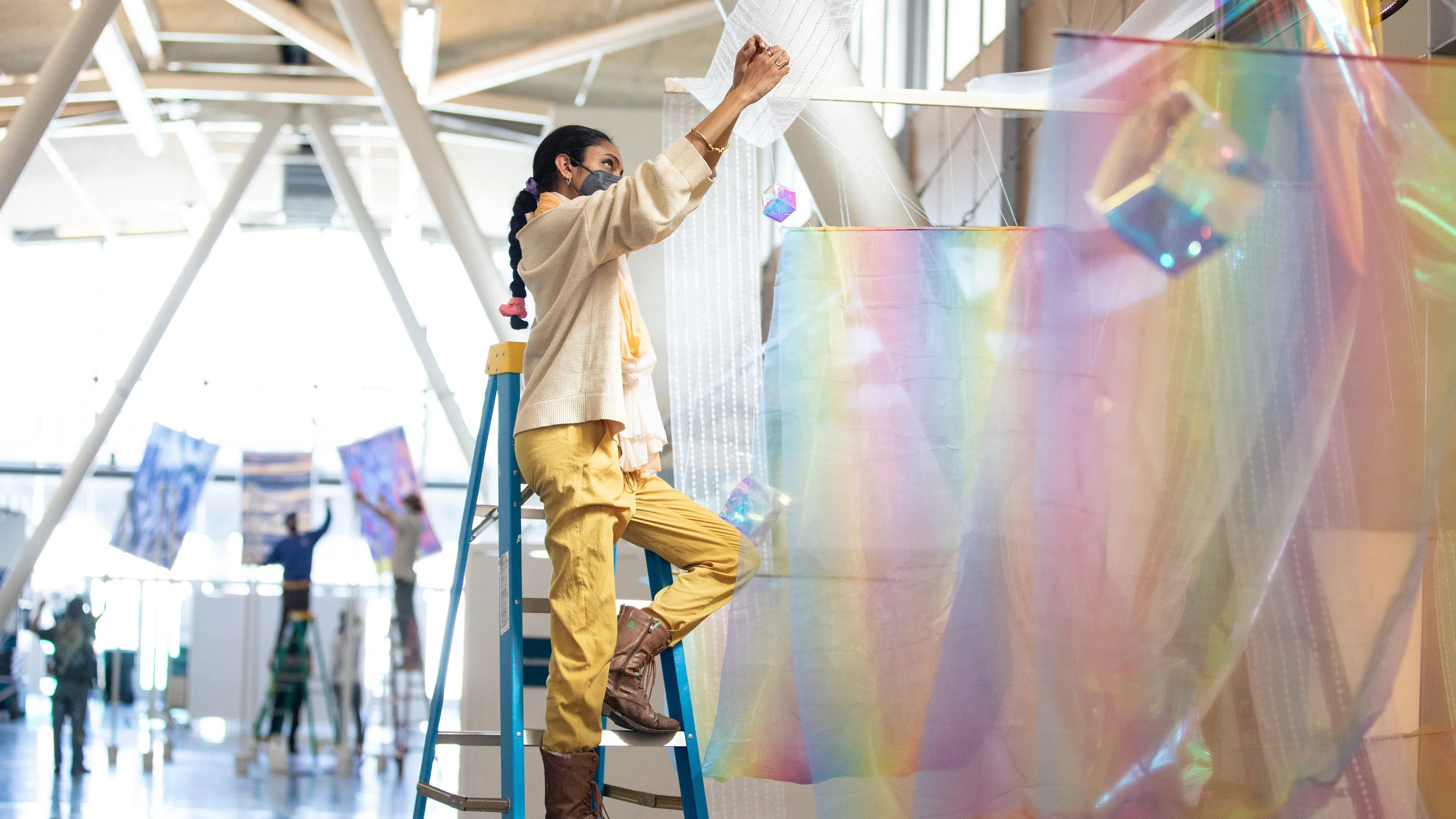 A person stands on a ladder, hanging an installation of gauzy mesh and prisms. Toward the back of the light-filled space, four people suspend large-scale photographs.