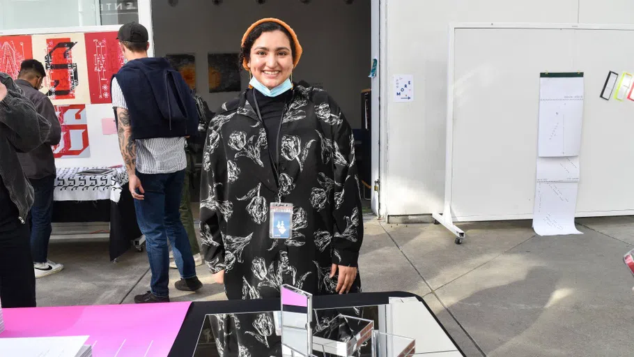 A student poses in front of their publication on a mirrored table at the MFA in Design Book Fair.