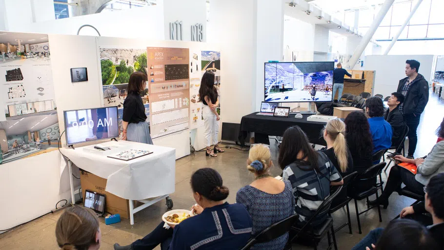 Two students stand in front of a crowd presenting work on the walls and a large monitor.