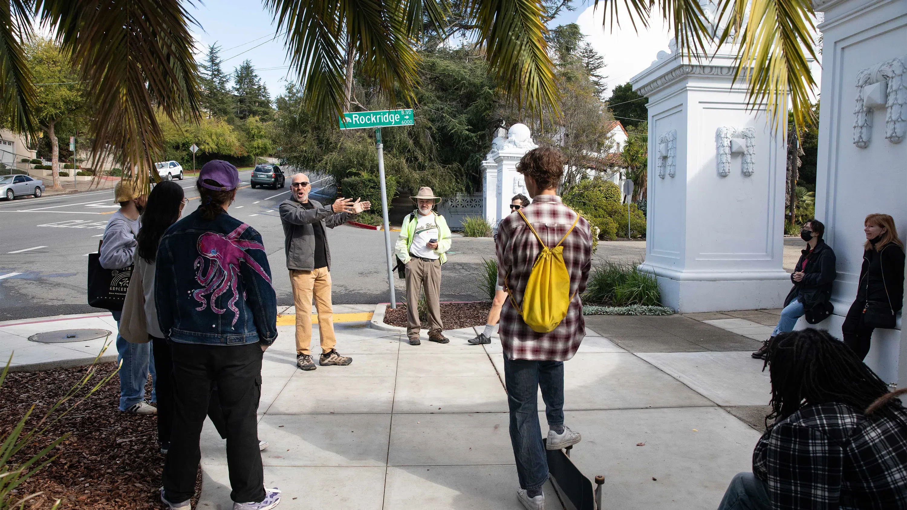A professor gestures and talks to a small group standing on the corner near a green street sign that says Rockridge.