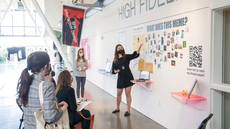 A student shares work during critique, gesturing toward a wall with images below vinyl reading”What does this Meme?”