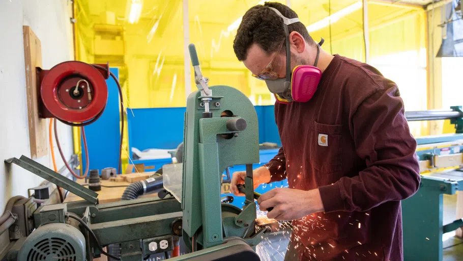 A student uses a bandsaw in the Metal Shop, with sparks flying dramatically around him.