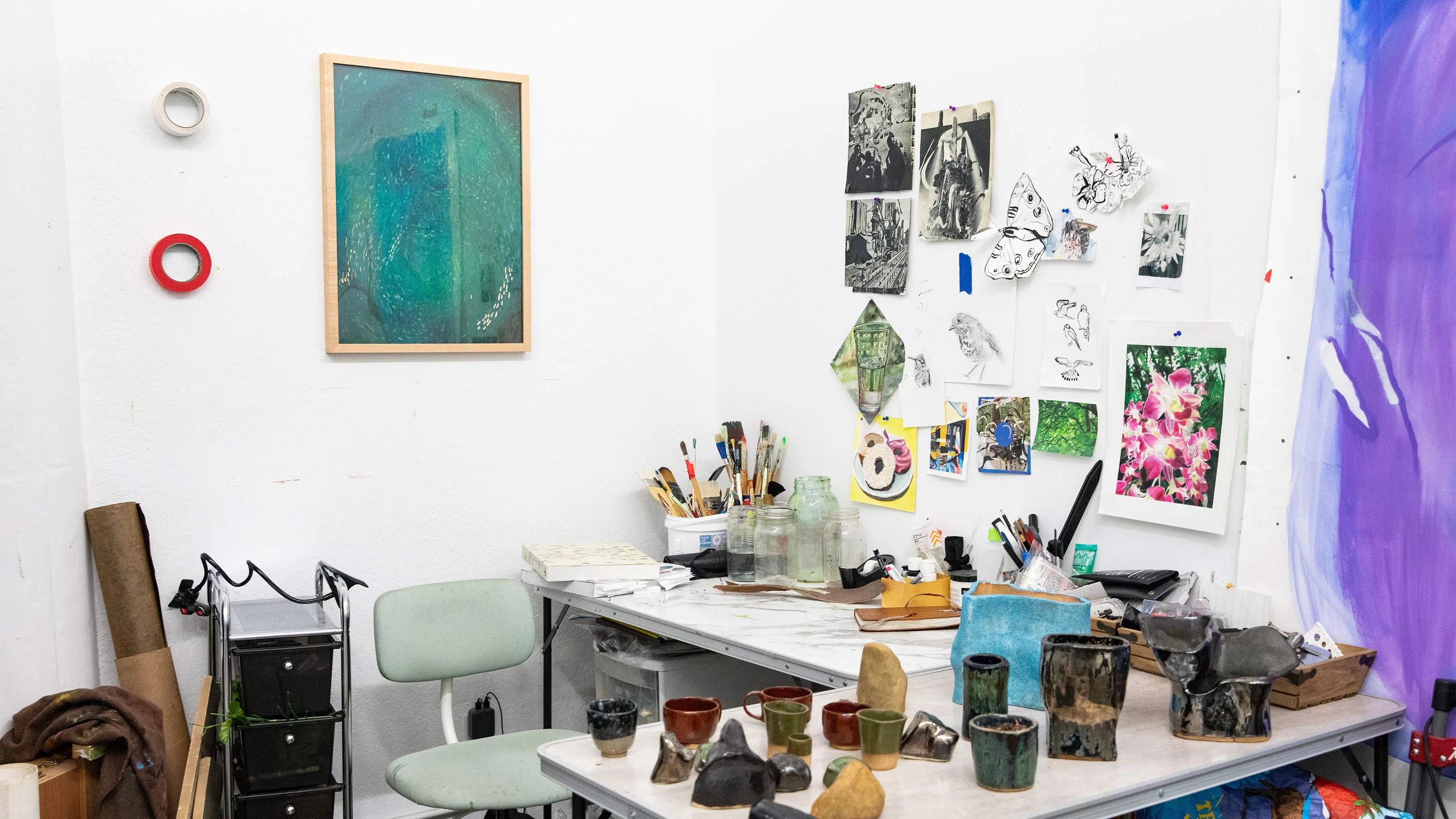 A corner of a grad student’s studio space that has art work pinned to the walls, a chair, L-shaped desk lined with paintbrushes and ceramics.